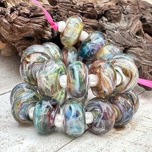 May include: A collection of round glass beads featuring swirling patterns of blue, green, brown, and gold glitter, separated by small white spacer beads on a bright pink string. The beads have a glossy finish and varied color distributions.