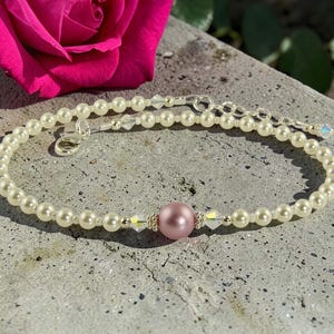 May include: A pearl bracelet with a central pink bead and crystal accents. The bracelet is made of small, creamy-white pearls and has a silver clasp. A bright pink rose is in the background. The bracelet's length is adjustable.