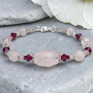 May include: A beaded bracelet featuring a large, faceted, pale pink rose quartz center bead flanked by deep red bicone crystals and smaller round rose quartz beads. The design includes small silver-toned metal spacers and a silver-toned lobster claw clasp.