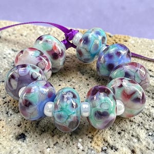 May include: A handmade bracelet featuring ten colorful glass beads. The beads are a mix of purple, blue, green, and red, with a marbled effect. The bracelet is strung on a purple cord and is a unique piece of jewelry.