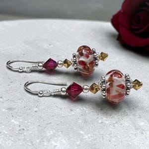 May include: A pair of dangle earrings featuring silver-tone hooks, faceted deep pink bicone crystals, small metallic spacers, and round lampwork glass beads with a white and red marbled pattern, finished with a small gold-tone pointed bead.