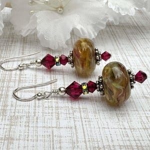 May include: A pair of handcrafted earrings featuring silver-tone hooks, maroon crystal accents, and oval-shaped, marbled glass beads in shades of brown and green. The earrings are set against a white and gray wood-grain surface.
