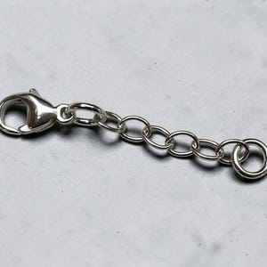 May include: A silver-colored jewelry extender with a lobster clasp on one end and a jump ring on the other. The extender chain is composed of small, oval-shaped links. The clasp and jump ring are also silver-toned.