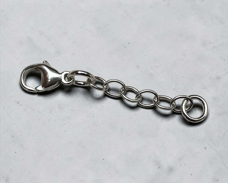 May include: A silver-colored jewelry extender with a lobster clasp on one end and a jump ring on the other. The extender chain is composed of small, oval-shaped links. The clasp and jump ring are also silver-toned.