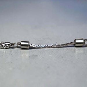 May include: A silver-toned jewelry extender chain featuring a flat, woven-style link design. The extender includes a polished lobster claw clasp on one end and a small circular jump ring on the other, with smooth cylindrical metal end caps.