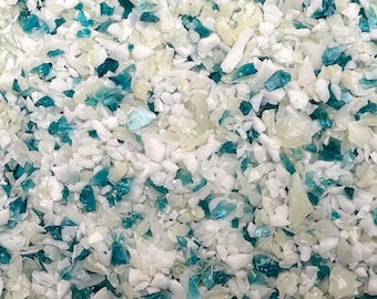 May include: Close-up of a pile of crushed glass pieces. The pieces are a mix of white, light blue, and clear, creating a textured, mosaic-like appearance. The fragments vary in size and shape, with some appearing translucent.