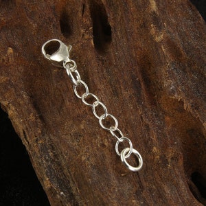 May include: A silver-colored chain extender with a lobster clasp and several small, round links. The extender is designed to lengthen necklaces or bracelets. The background is a piece of dark brown wood.