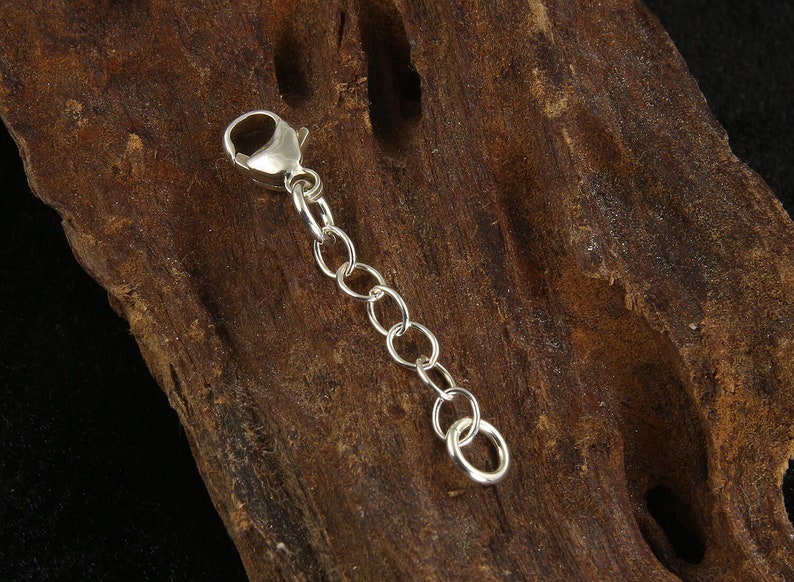 May include: A silver-colored chain extender with a lobster clasp and several small, round links. The extender is designed to lengthen necklaces or bracelets. The background is a piece of dark brown wood.