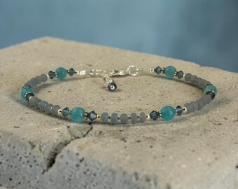 May include: A beaded bracelet featuring gray and teal beads, interspersed with small silver accents. The bracelet has a delicate chain and clasp. The teal beads are larger and more prominent, adding a pop of color to the design.