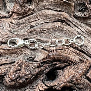 May include: A silver-colored necklace extender with a lobster clasp and chain links. The extender is designed to add length to necklaces or bracelets. The background is a textured, brown wooden surface.