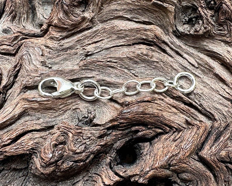 May include: A silver-colored necklace extender with a lobster clasp and chain links. The extender is designed to add length to necklaces or bracelets. The background is a textured, brown wooden surface.