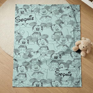 Puede incluir: Una manta de beb&eacute; azul claro con un patr&oacute;n repetido de bocetos de Mickey Mouse en negro. La manta tiene el nombre "Sophie" escrito en escritura negra. Un peque&ntilde;o oso de peluche est&aacute; al lado de la manta.