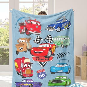 Puede incluir: Una manta azul claro con ilustraciones de coches de dibujos animados, incluyendo un cami&oacute;n de bomberos rojo, un Fabulous Hudson Hornet azul y un coche de carreras rojo llamado David, con se&ntilde;alizaci&oacute;n de la Ruta 66. Marcado "Enviado desde EE. UU."