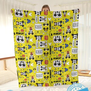 Puede incluir: Una manta amarilla con un patr&oacute;n repetido de ilustraciones de Mickey Mouse. La manta incluye el nombre "ROBERT" en cajas y el texto "Custom name". La manta se muestra sobre una cama.