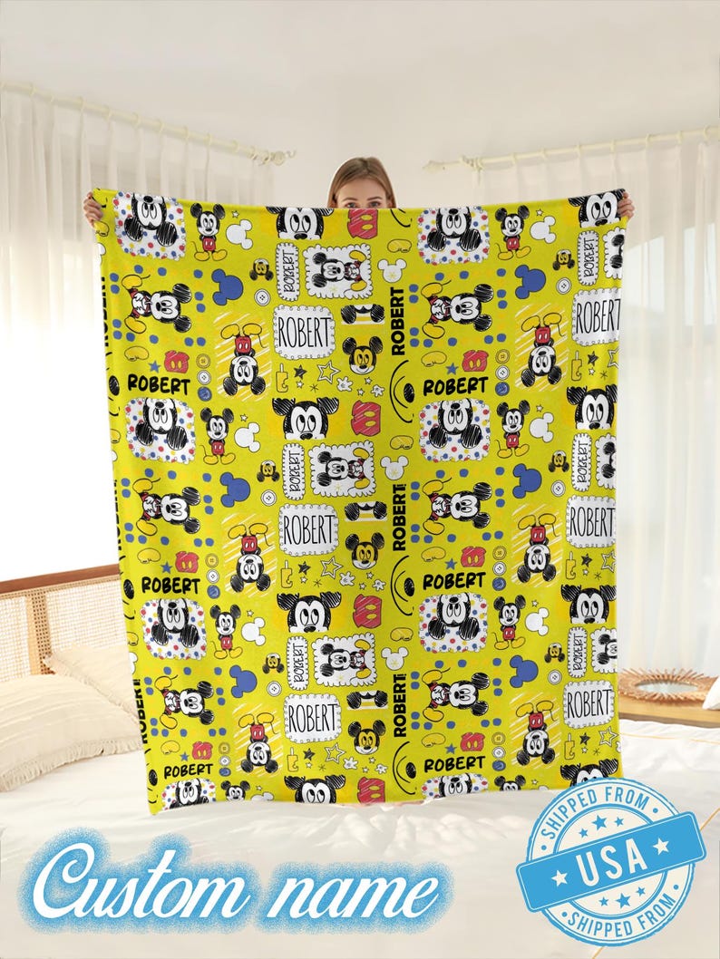 Puede incluir: Una manta amarilla con un patr&oacute;n repetido de ilustraciones de Mickey Mouse. La manta incluye el nombre "ROBERT" en cajas y el texto "Custom name". La manta se muestra sobre una cama.