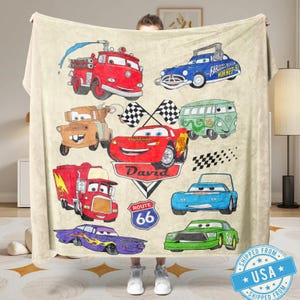 Puede incluir: Una manta beige con ilustraciones de coches de dibujos animados en rojo, azul, verde y morado. La manta incluye un cami&oacute;n de bomberos rojo, un coche de carreras azul y una furgoneta verde. Tambi&eacute;n tiene el nombre "David" y un letrero de la Ruta 66.