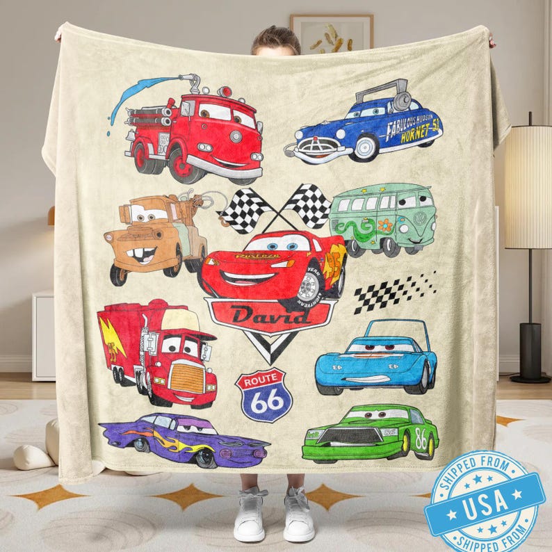 Puede incluir: Una manta beige con ilustraciones de coches de dibujos animados en rojo, azul, verde y morado. La manta incluye un cami&oacute;n de bomberos rojo, un coche de carreras azul y una furgoneta verde. Tambi&eacute;n tiene el nombre "David" y un letrero de la Ruta 66.