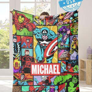 May include: A colorful blanket featuring a collage of comic book characters, including Captain America, Iron Man, and the Hulk. The blanket has a red and white rectangle with the name "MICHAEL" printed on it. The blanket is being held up by a person.