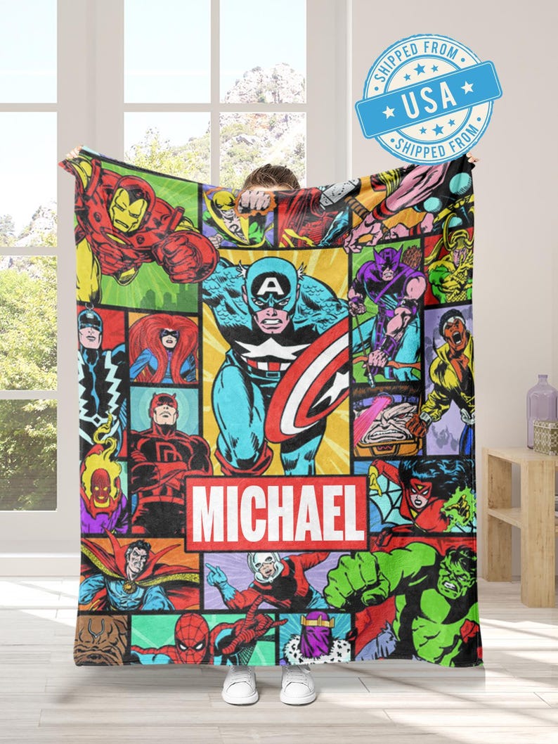 May include: A colorful blanket featuring a collage of comic book characters, including Captain America, Iron Man, and the Hulk. The blanket has a red and white rectangle with the name "MICHAEL" printed on it. The blanket is being held up by a person.