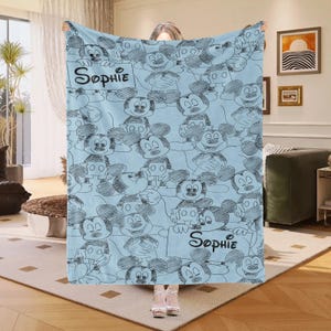 Puede incluir: Una manta azul claro con un patr&oacute;n repetido de bocetos de Mickey Mouse en negro. La manta tiene el nombre "Sophie" escrito en escritura negra en dos letreros. La manta est&aacute; hecha de un material suave.