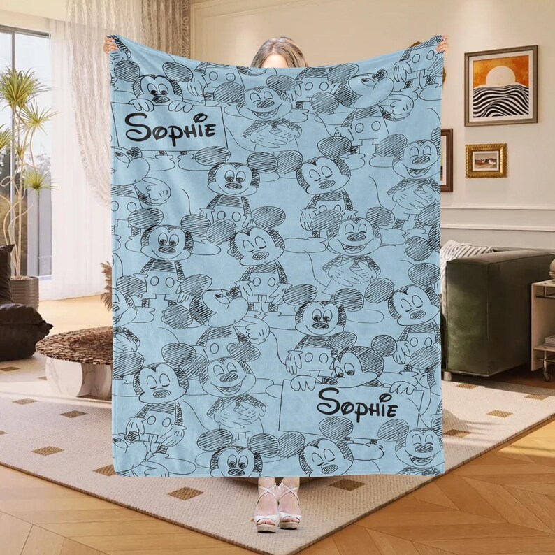 Puede incluir: Una manta azul claro con un patr&oacute;n repetido de bocetos de Mickey Mouse en negro. La manta tiene el nombre "Sophie" escrito en escritura negra en dos letreros. La manta est&aacute; hecha de un material suave.