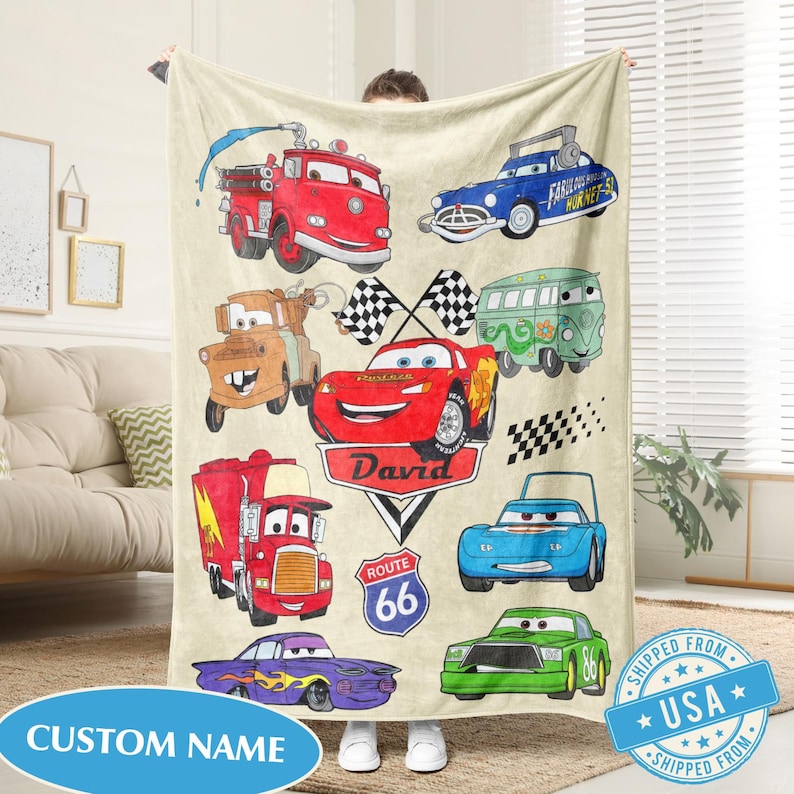 Puede incluir: Una manta de forro polar beige con dibujos de coches de dibujos animados en varios colores. La manta incluye un cami&oacute;n de bomberos rojo, un coche azul con el texto "Fabulous Hornets" y un coche de carreras rojo con el nombre "David". Tambi&eacute;n tiene un letrero de "Ruta 66" y una etiqueta de "Custom Name".