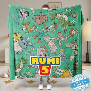May include: A green blanket featuring cartoon characters from the Toy Story franchise. The blanket has a repeating pattern of characters like Woody, Buzz Lightyear, and Rex. The name "RUMI" and the number "5" are prominently displayed in a logo.