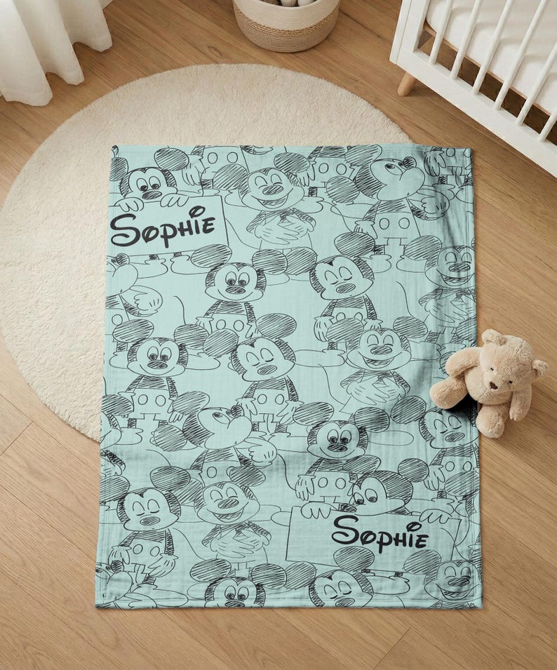 Puede incluir: Una manta de beb&eacute; azul claro con un patr&oacute;n repetido de bocetos de Mickey Mouse en negro. La manta tiene el nombre "Sophie" escrito en escritura negra. Un peque&ntilde;o oso de peluche est&aacute; al lado de la manta.