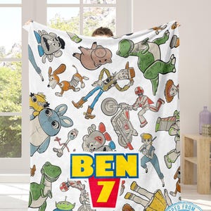 May include: White blanket featuring colorful cartoon characters from the Toy Story franchise. The blanket has a repeating pattern of characters and the name "BEN 7" in yellow and red. The blanket is made of a soft material.