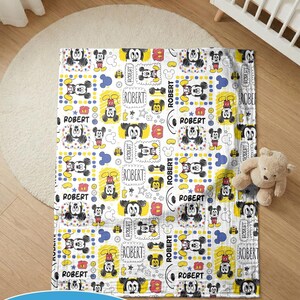 Puede incluir: Una manta de beb&eacute; blanca con un patr&oacute;n repetido de ilustraciones de Mickey Mouse, c&iacute;rculos amarillos y el nombre "ROBERT" en un marco rectangular. La manta est&aacute; sobre un suelo de madera.