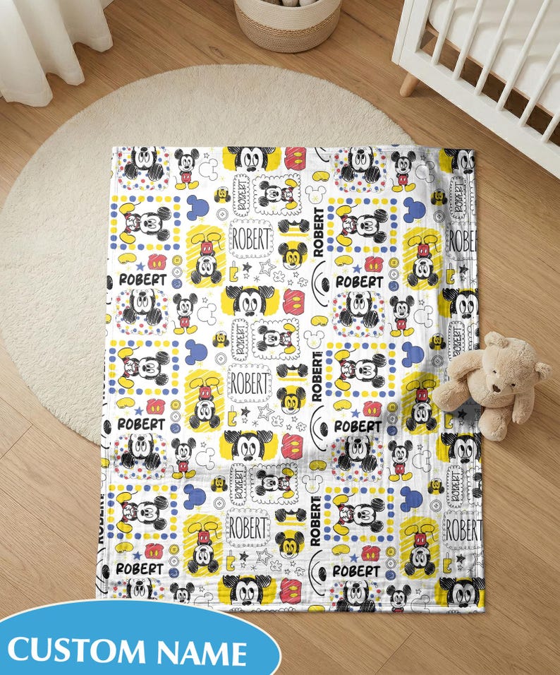 Puede incluir: Una manta de beb&eacute; blanca con un patr&oacute;n repetido de ilustraciones de Mickey Mouse, c&iacute;rculos amarillos y el nombre "ROBERT" en un marco rectangular. La manta est&aacute; sobre un suelo de madera.