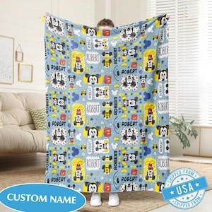 Puede incluir: Una manta azul claro con un patr&oacute;n repetido de ilustraciones de Mickey Mouse, estrellas y el nombre "ROBERT" en un marco rectangular. La manta tiene una etiqueta "CUSTOM NAME".