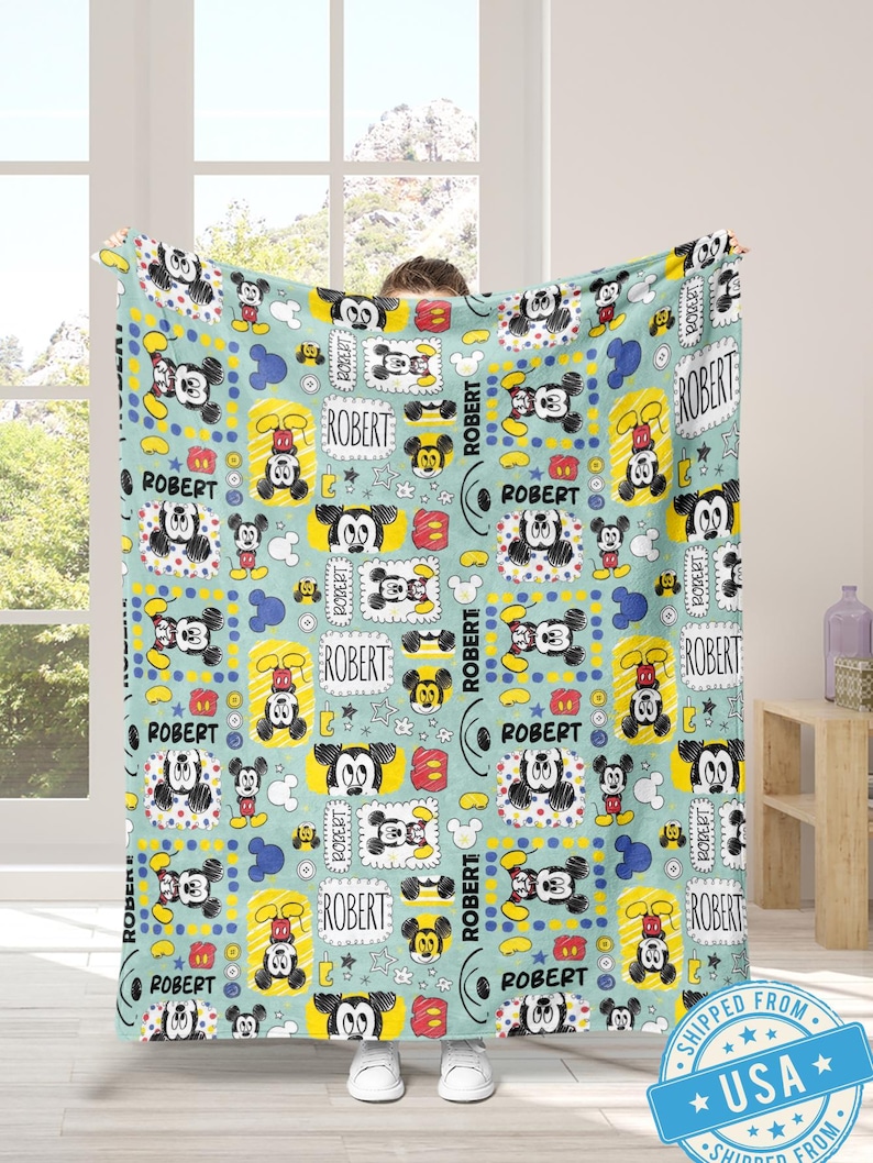 Puede incluir: Una manta azul claro con un patr&oacute;n repetido de ilustraciones de Mickey Mouse y el nombre "ROBERT" en un marco rectangular. El dise&ntilde;o incluye acentos amarillos, rojos y azules, creando una est&eacute;tica l&uacute;dica y colorida.