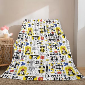 Puede incluir: Manta blanca con un patr&oacute;n repetido de ilustraciones de Mickey Mouse en negro, rojo, amarillo y azul. La manta tiene el nombre "ROBERT" impreso. La manta est&aacute; sobre una superficie.