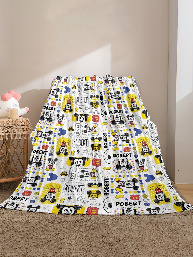 Puede incluir: Manta blanca con un patr&oacute;n repetido de ilustraciones de Mickey Mouse en negro, rojo, amarillo y azul. La manta tiene el nombre "ROBERT" impreso. La manta est&aacute; sobre una superficie.
