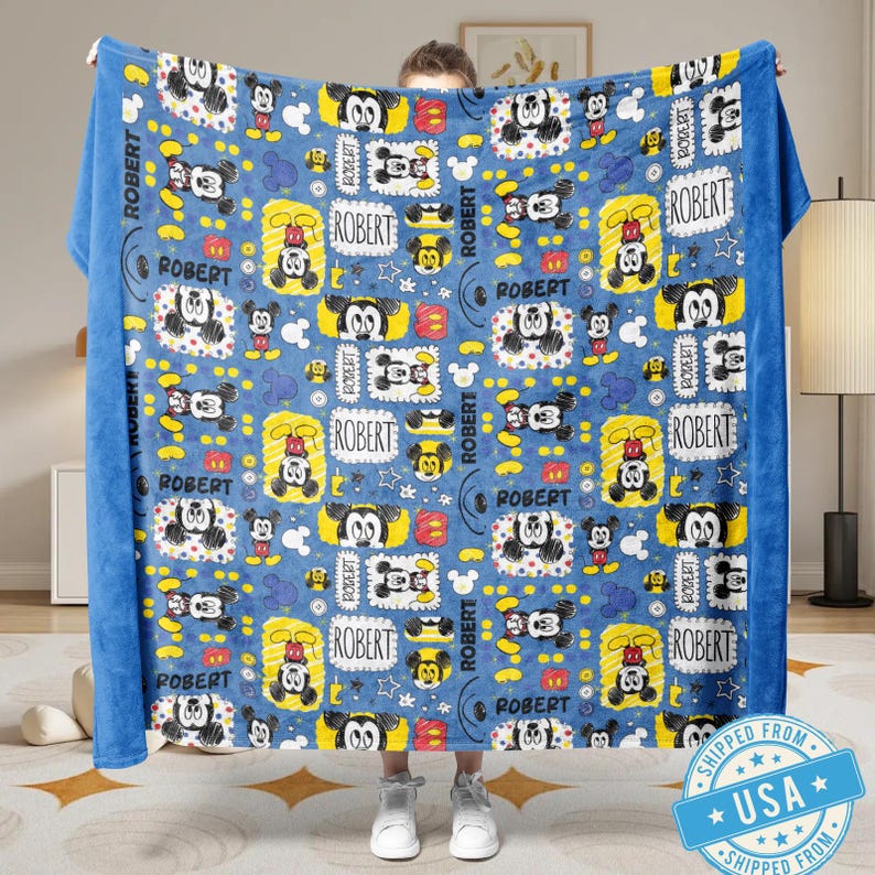 Puede incluir: Una manta azul con un patr&oacute;n repetido de personajes de Mickey Mouse en varias poses y dise&ntilde;os. La manta incluye el nombre "ROBERT" en un patr&oacute;n repetido. El borde es de color azul liso.