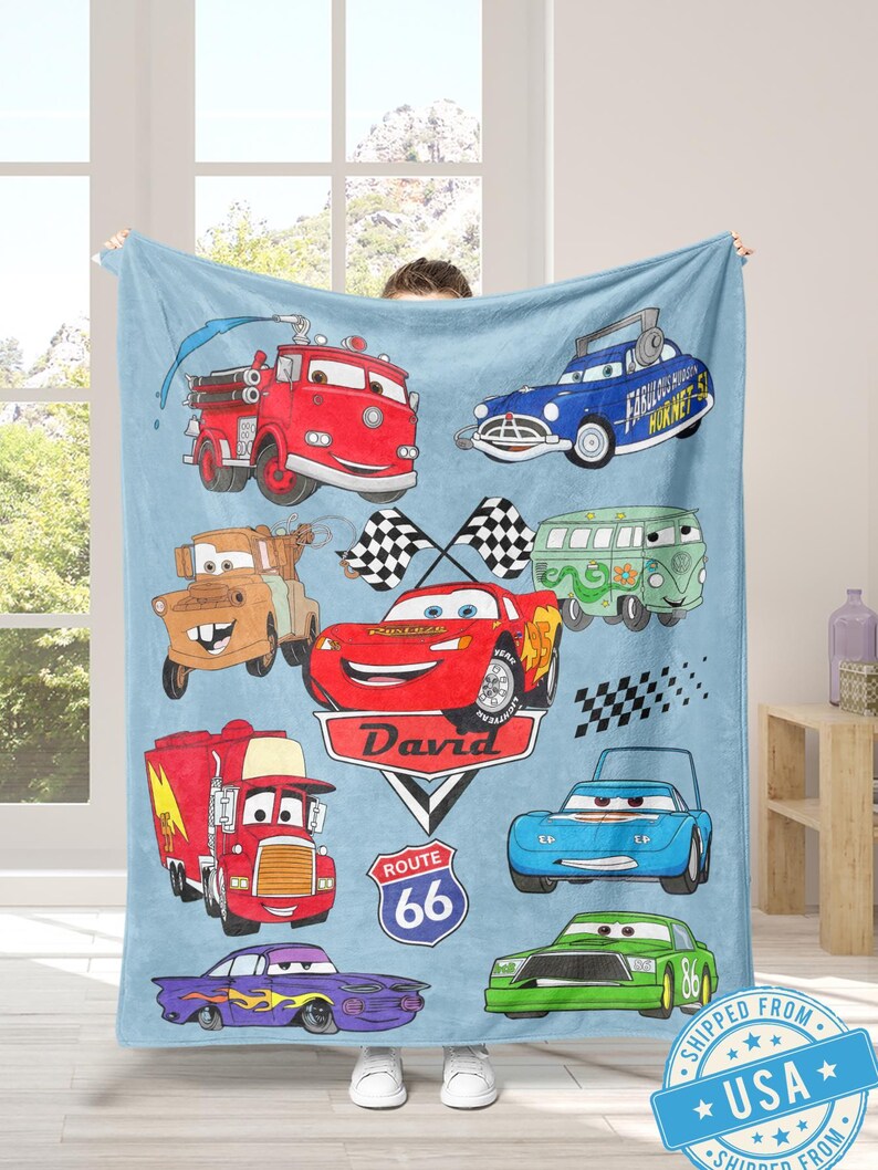 Puede incluir: Una manta azul claro con ilustraciones de coches de dibujos animados. La manta muestra varios dise&ntilde;os de coches, incluyendo un cami&oacute;n de bomberos rojo, un coche azul con el texto "Fabulous Hudson Hornet" y un coche de carreras rojo con el nombre "David". Incluye un letrero de la Ruta 66.