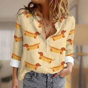May include: A button-down shirt with a repeating pattern of dachshunds in hot dog buns. The shirt is a light beige color with long sleeves and white cuffs. The dachshunds are brown with yellow buns.
