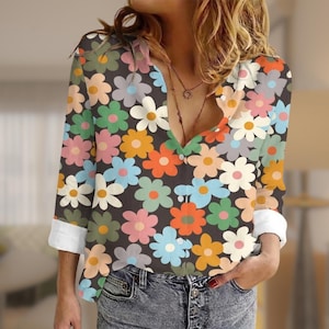 May include: A long-sleeved floral print shirt with a deep V-neck. The shirt features a pattern of colorful flowers in shades of pink, orange, blue, and green on a dark background. The cuffs are white, and the shirt is paired with blue jeans.