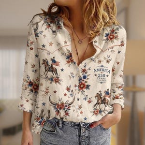 May include: A long-sleeved button-down shirt in an off-white color featuring a repeating print of horses, American flags, stars, horseshoes, and floral patterns. The text AMERICA 250 YEARS 1776-2026 is printed on the front right side.