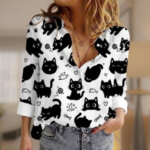 May include: A white long-sleeve button-down shirt featuring a repeating pattern of black cats, small hearts, yarn balls, crescent moons, and fish bones. The shirt has a collar, a front button placket, and sleeves that are rolled up at the cuffs.