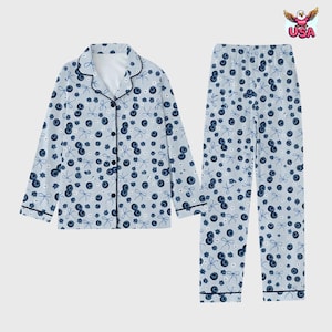 May include: Light blue pajama set featuring a pattern of blueberries and bows. The long-sleeved top has a button-down front with black trim, and the pants have an elastic waistband. The set is designed for comfort and relaxation.