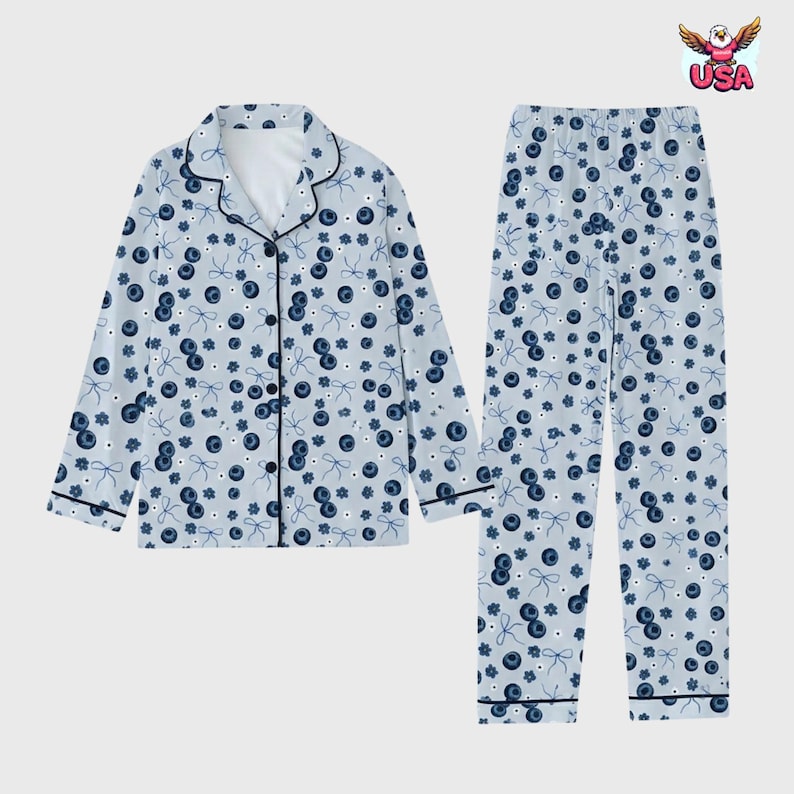 May include: Light blue pajama set featuring a pattern of blueberries and bows. The long-sleeved top has a button-down front with black trim, and the pants have an elastic waistband. The set is designed for comfort and relaxation.