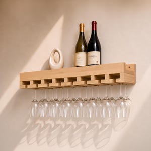 Puede incluir: Un botellero de pared de madera clara con un estante superior que sostiene dos botellas de vino y un objeto ovalado decorativo. El estante incluye ranuras integradas en la parte inferior para colgar varias copas de vino de cristal transparente.