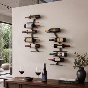 May include: Two black metal wall-mounted wine racks holding multiple wine bottles. The racks feature a curved vertical spine with horizontal shelves. Below, a wooden console table holds two wine glasses, a bottle, and a black vase.