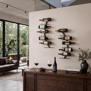 May include: A modern living room scene featuring two wall-mounted wine racks holding several wine bottles. A wooden sideboard displays wine glasses, a bottle of wine, and decorative items. A leather sofa and large windows complete the elegant setting.