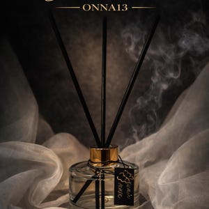 Könnte beinhalten: Ein klarer Glasdiffusor mit schwarzen Stäbchen und einem goldfarbenen Kragen. Auf dem Etikett steht "Kumin Musk". Die Worte "Black Water" sind in goldener Schrift über dem Diffusor geschrieben. Der Hintergrund ist ein weicher, drapierter Stoff.