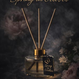 Puede incluir: Un difusor de vidrio transparente con una tapa dorada y varillas de madera. Una etiqueta negra con un diseño de mariposa y las palabras "Spring in October" cuelga de la botella. El fondo es oscuro con acentos florales.
