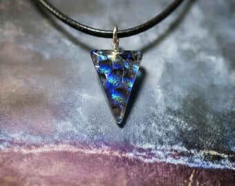 May include: A triangular pendant necklace with a black cord. The pendant is clear with blue and black iridescent inclusions. The pendant is suspended from a silver-colored bail. The background is a blurred mix of purple, blue, and brown.
