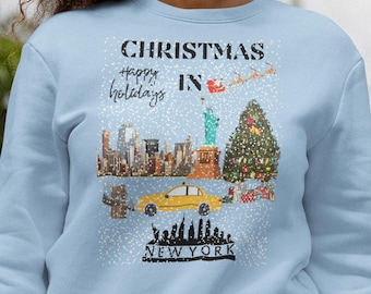 May include: A light blue crewneck sweatshirt featuring a graphic print of a snowy New York City scene with a yellow taxi, the Statue of Liberty, a Christmas tree, and the text CHRISTMAS Happy holidays IN NEW YORK with a flying Santa sleigh.
